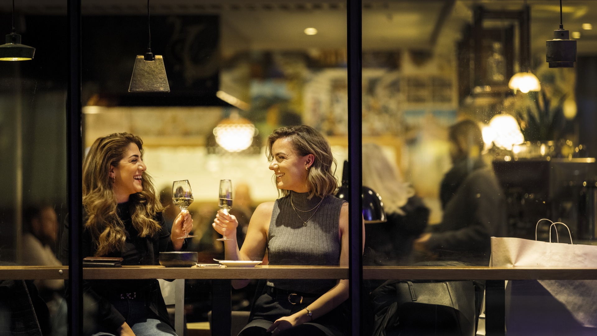 Two women are sitting at a table placed by a window. They each raise a glass toward each other for a toast.