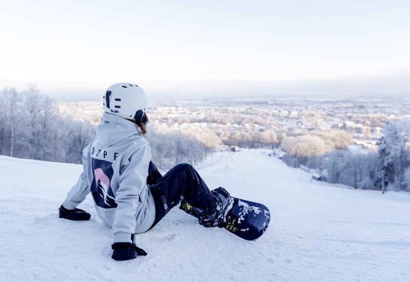 A girl sits at the top of the slalom hill looking at the view of Skövde. On her head she has a white helmet and black boots and a snowboard at her feet.