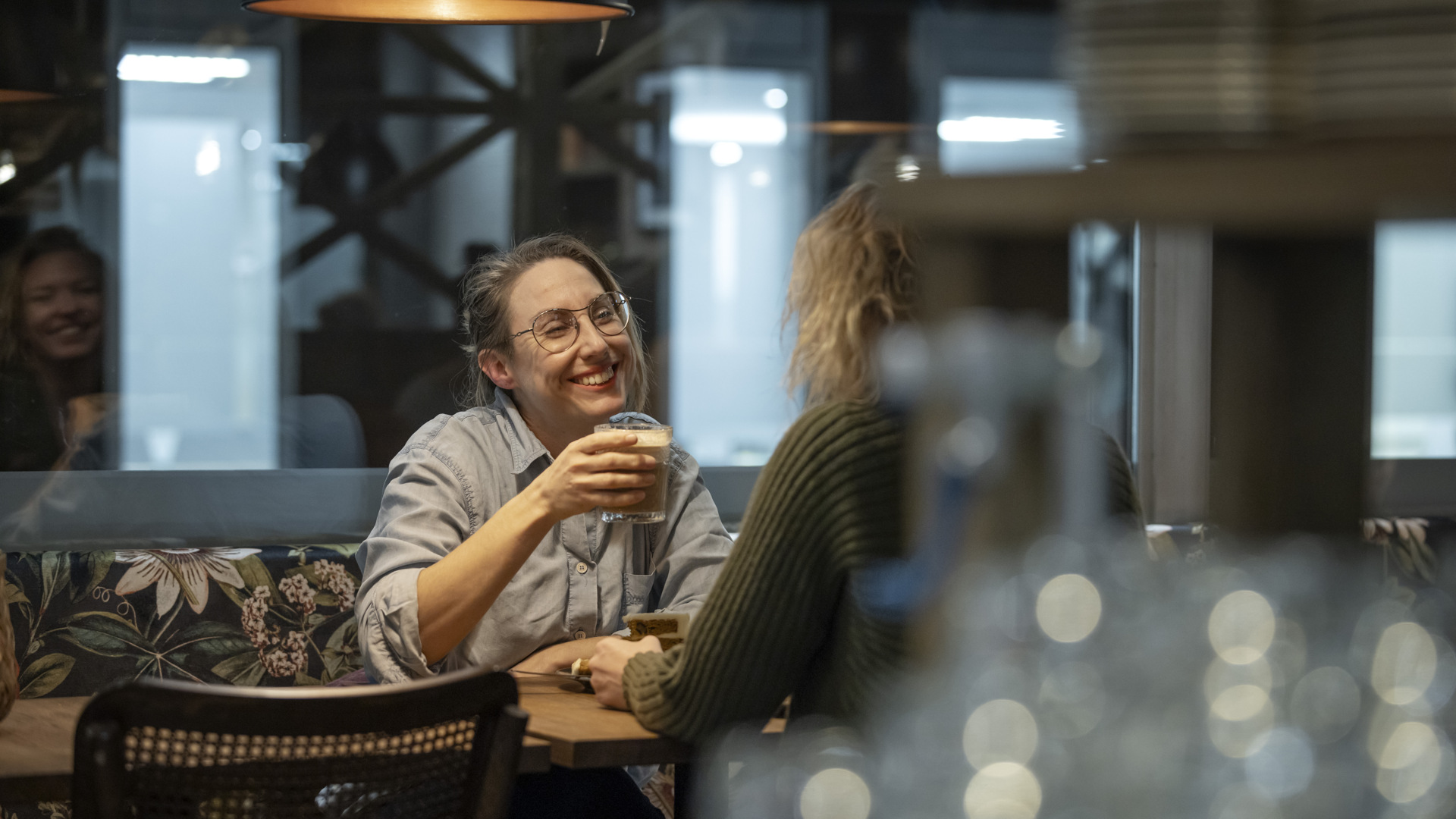 Two women are sitting at a table having coffee. One of them is smiling and holding a glass of coffee in her hand.