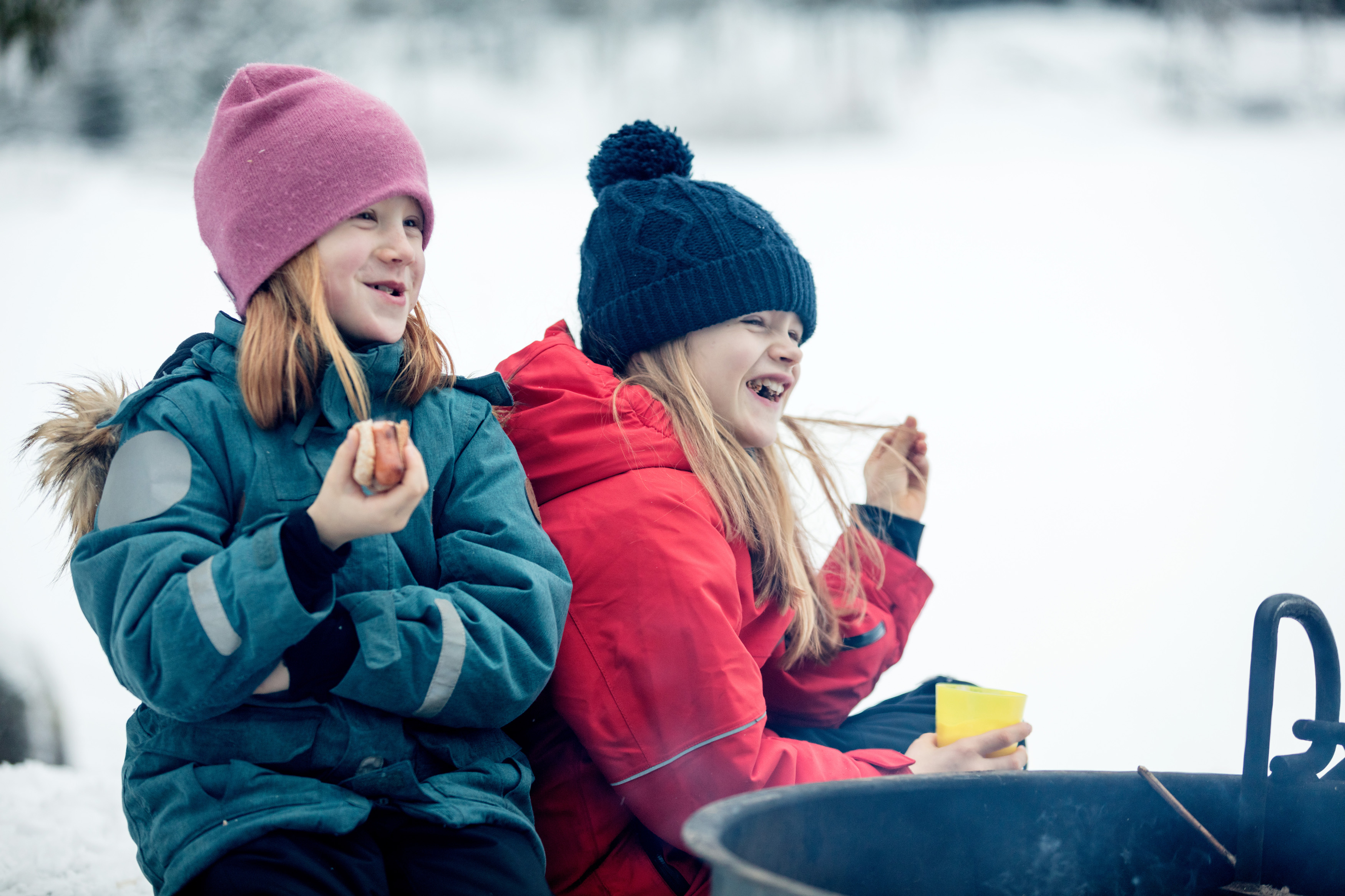 Family sitting by the fire surrounded by a winter landscape at Billingen moutain. Two girls are eating hot dogs and laughing.