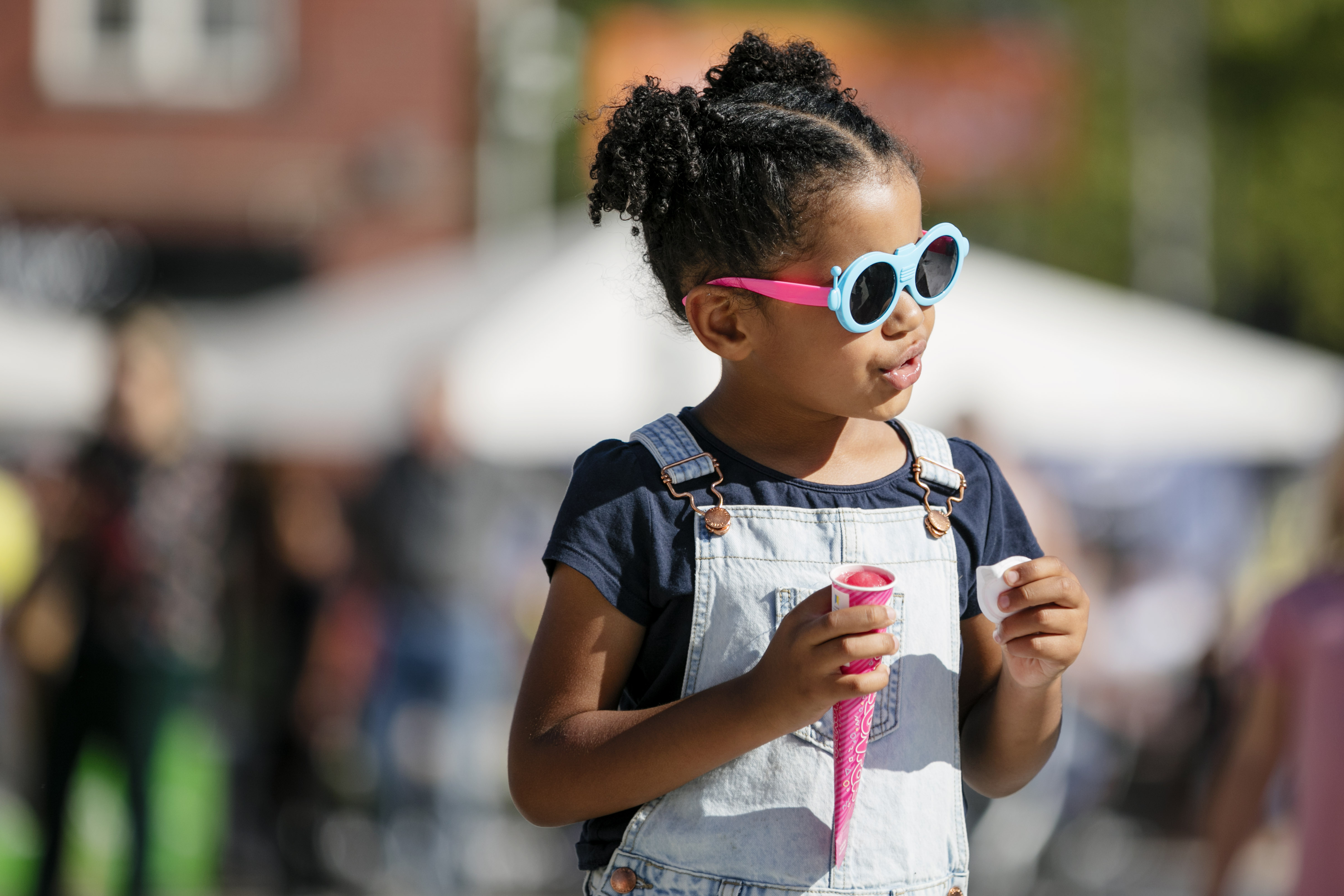 Little girl with black curly hair wearing a jeans outfit and cool looking glasses. In her hand she is holding a pink ice cream. 