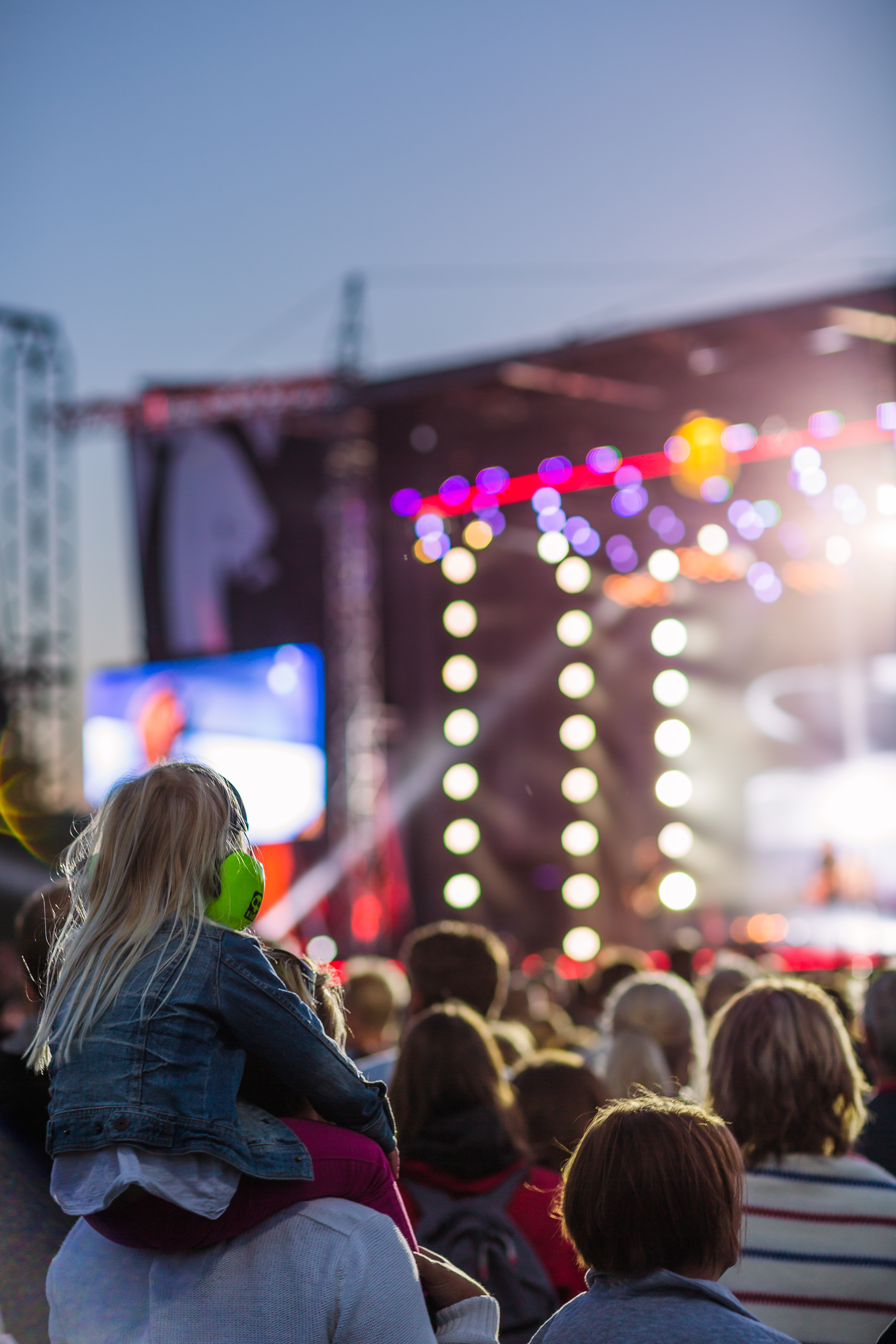 An ocean of people in front of the stage. In the foreground, a little girl sits on someone's shoulders with hearing aids on her head.