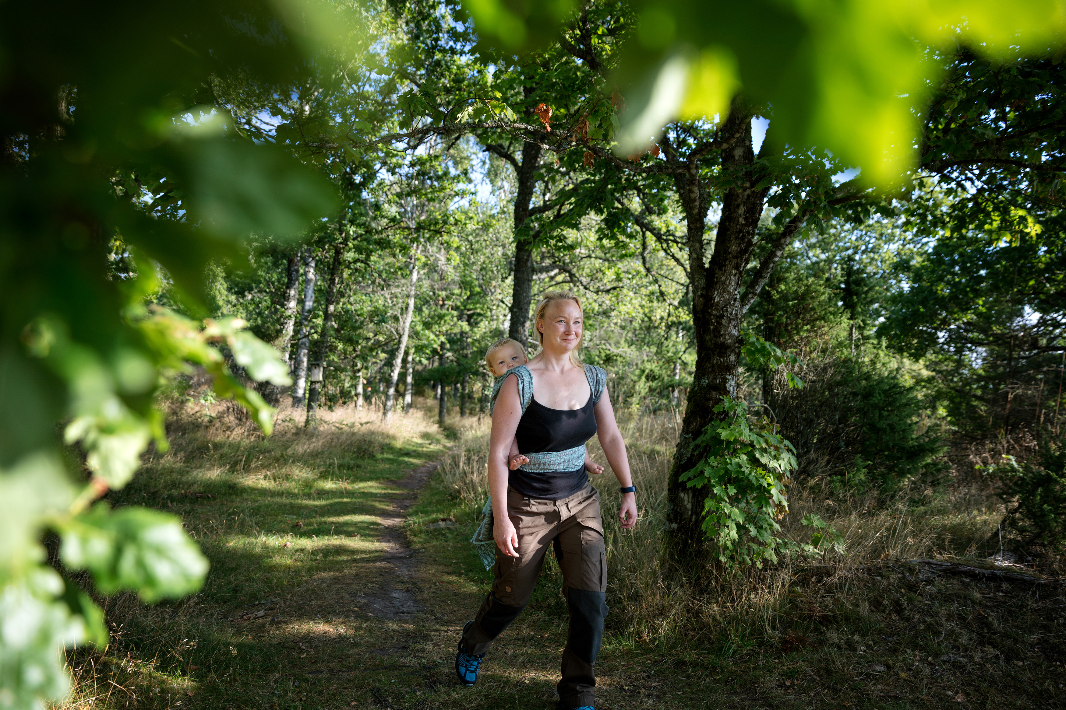 A woman with blonde hair and a baby wrap on her back is out walking in the green nature.