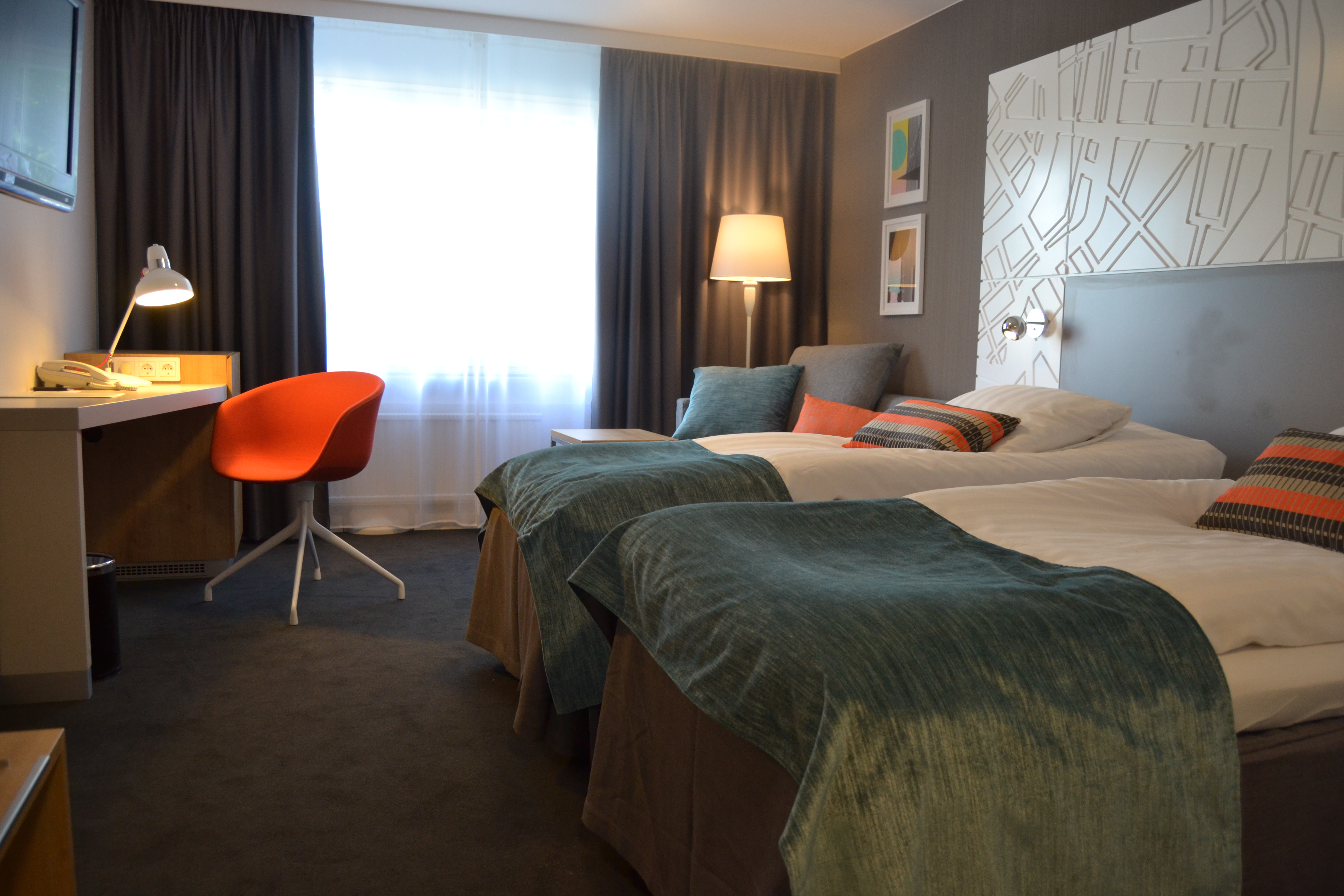 Two beds with turquoise bedspreads are standing next to each other in a hotel room. In front of the beds is also a desk with an orange chair.