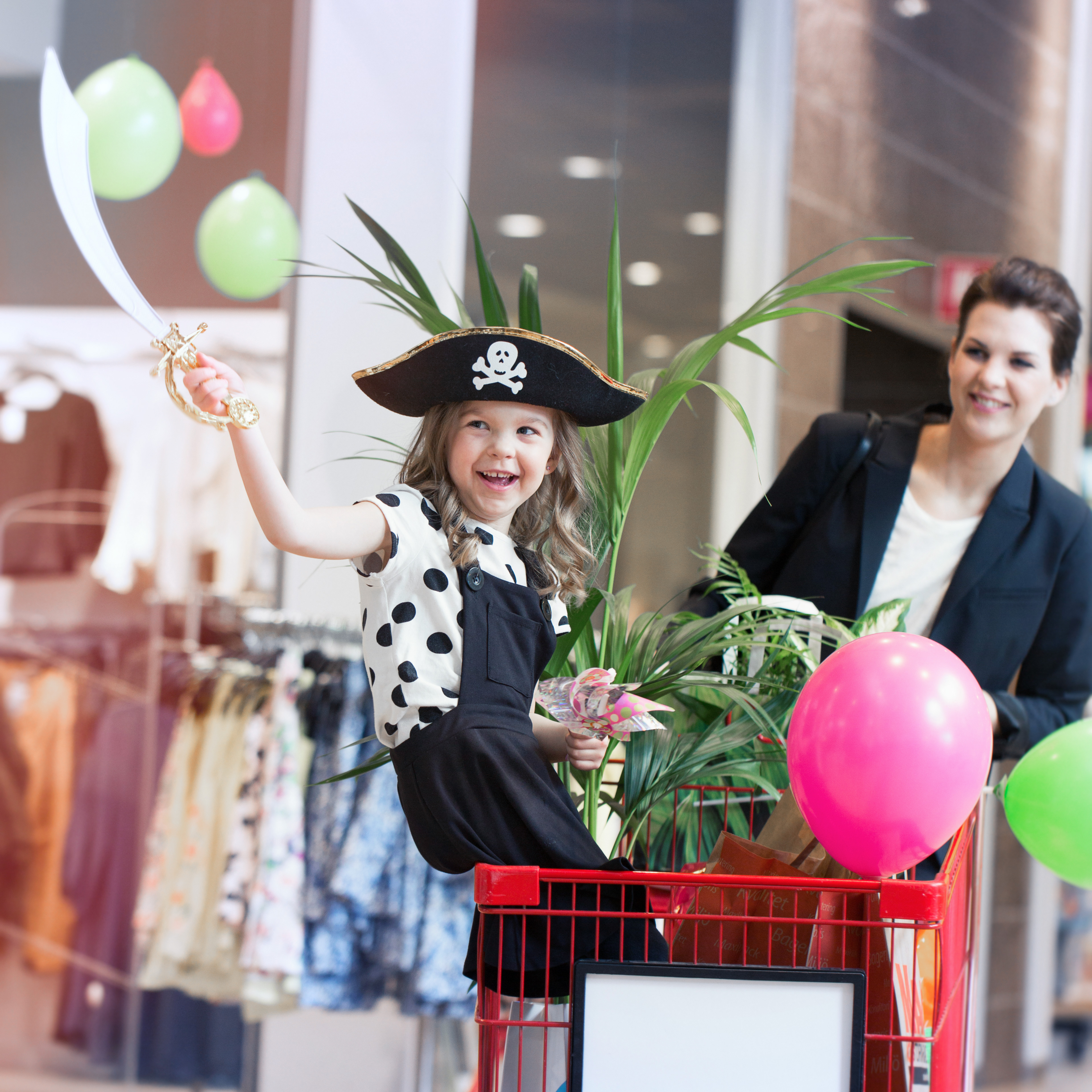 A little girl in pirate hat sits on the edge of a shopping cart and raises her pirate sword in the air. Behind, her mom drives the wagon full of flowers and balloons.