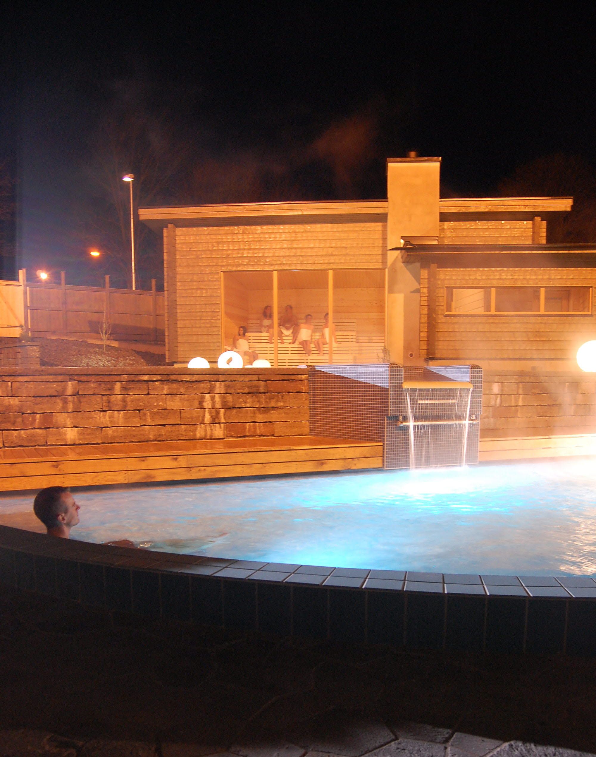 A man sits in a steaming outdoor pool. It is dark. In the background there is a wooden sauna with large windows.