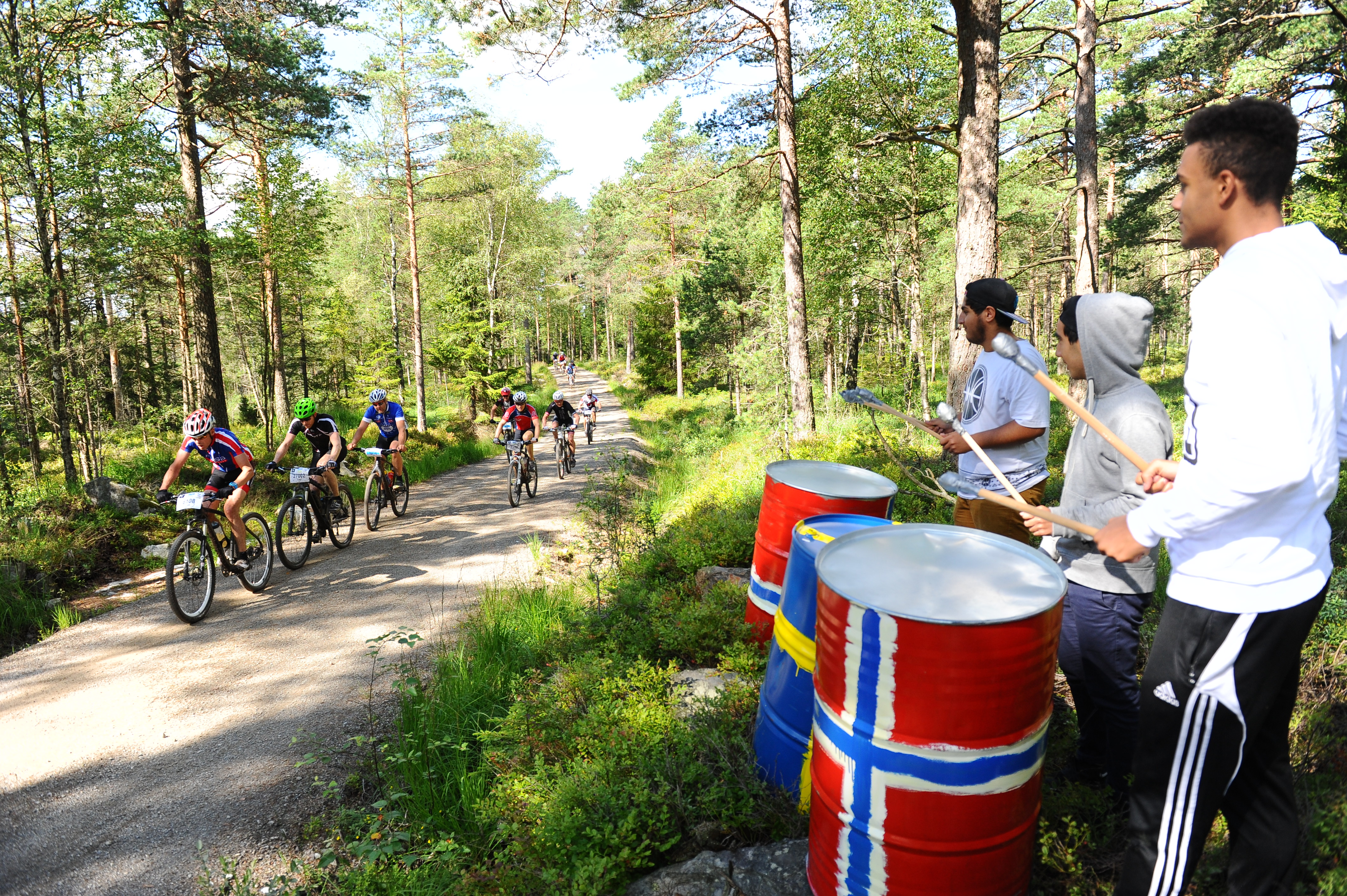 The cyclist race Grenserittet. The competitors are biking along a gravel road in the woods. Three spectators are standing on the side of the road, beating on drums. 