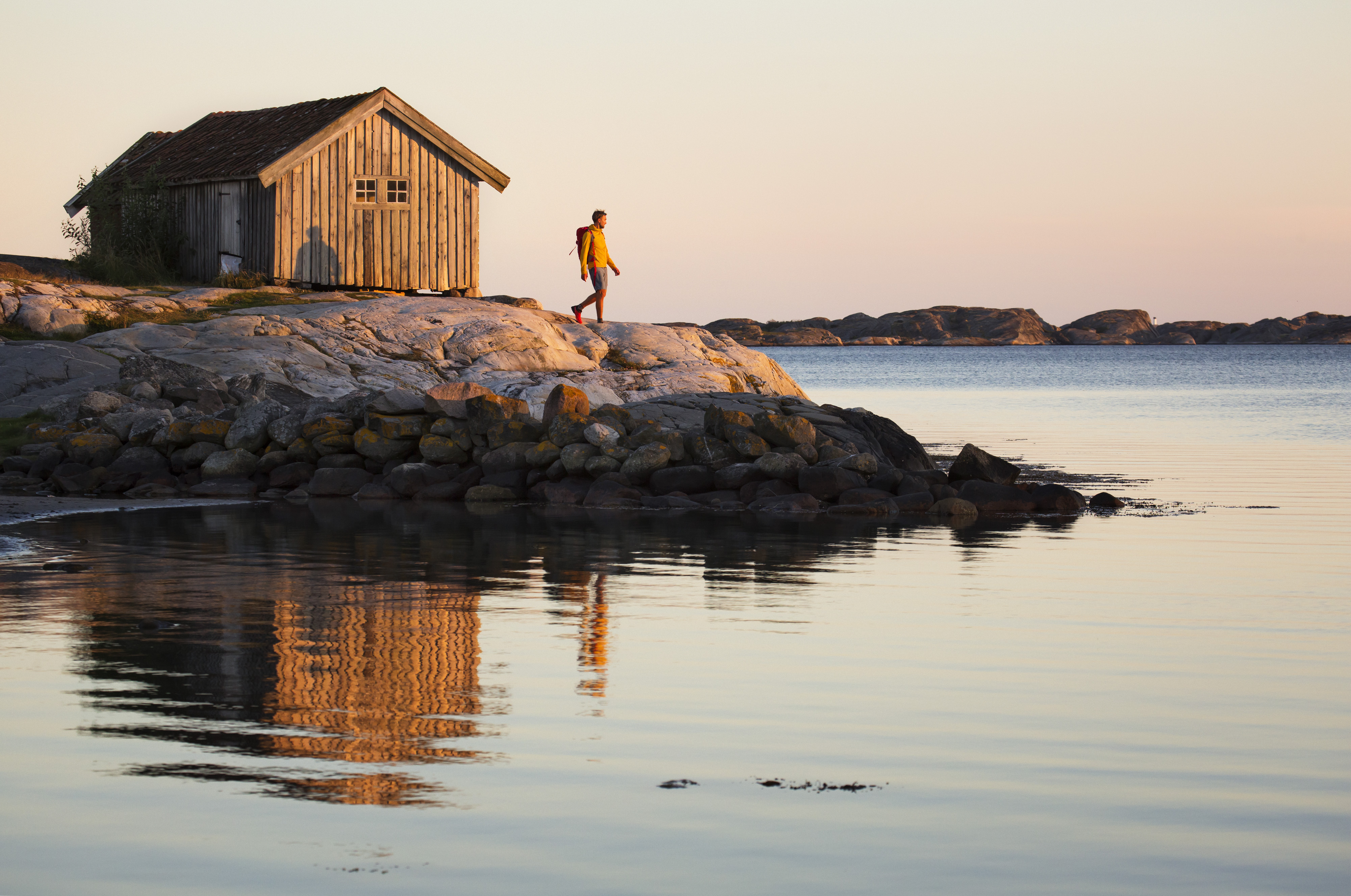 A person is standing on a cliff in front of a old and grey boathouse at South Koster. The ocean is calm and the sun is about to set. 