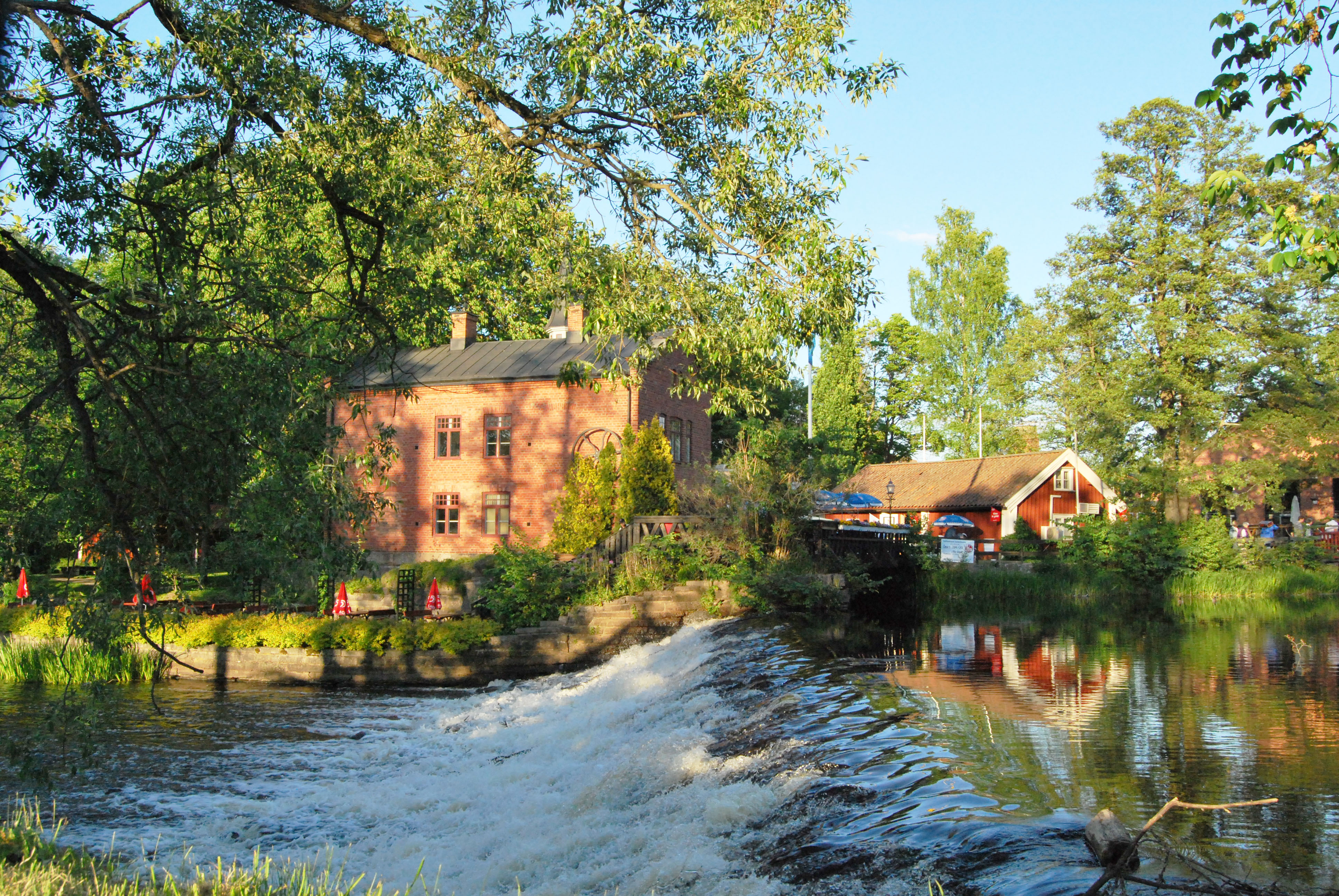 Picture of the Turbine House, which is Tidaholm Art Hall and Kaffestugan with a small waterfall in front of summer greenery.