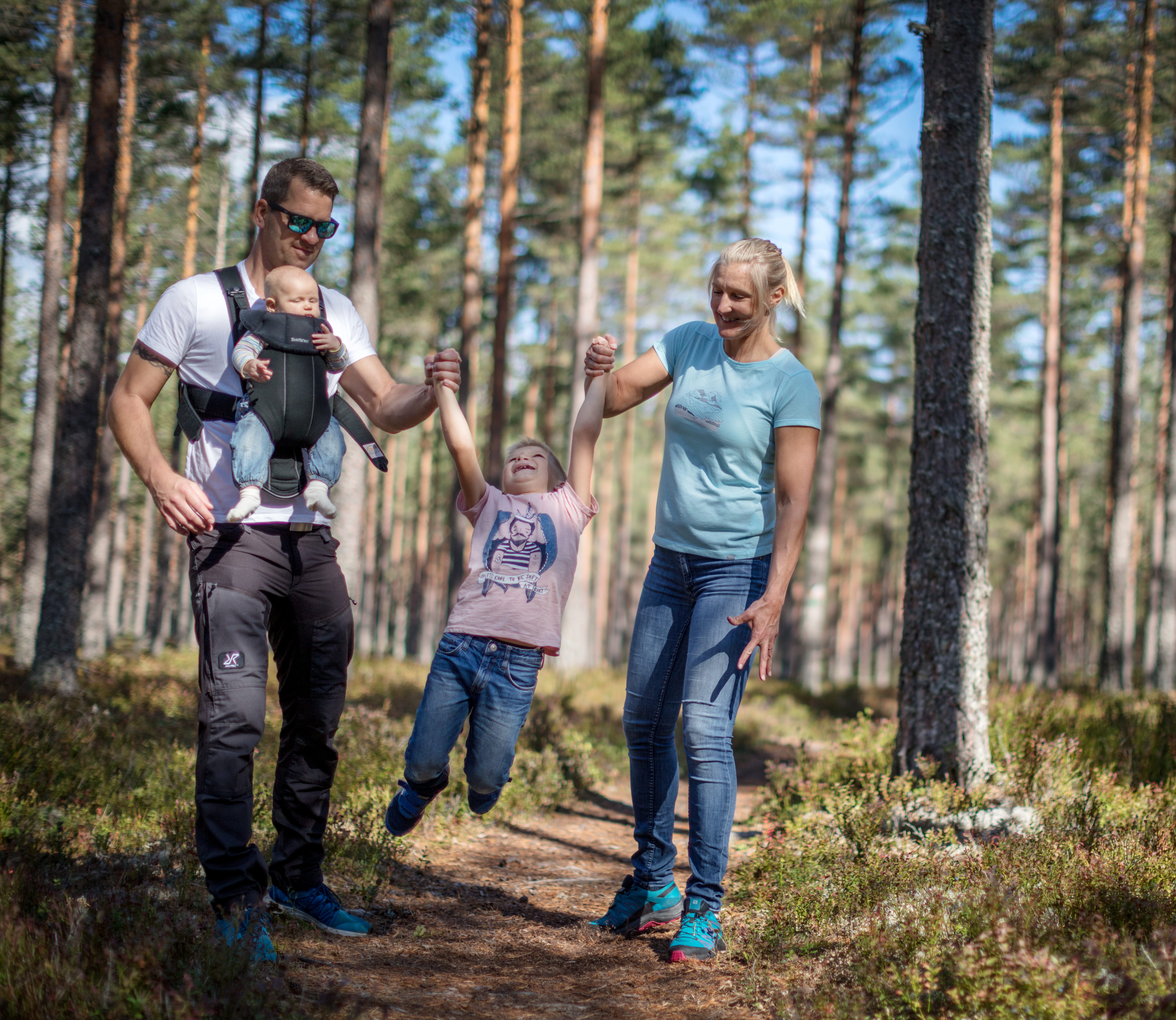 
A father and a mother hold their child between them and swing him back and forth. The father has an infant in a harness on his stomach