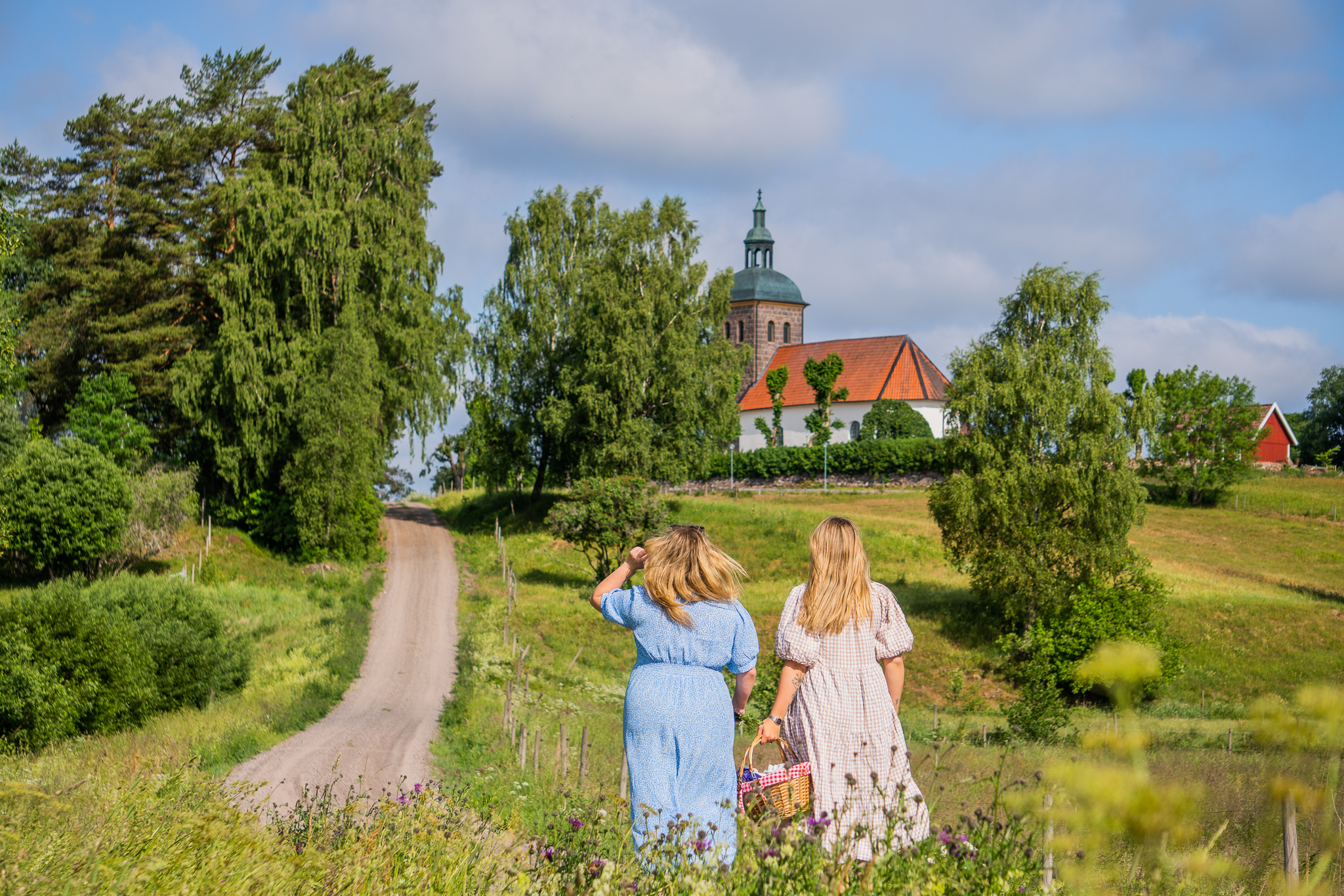 Two women dressed in summer dresses look out across a field and a church.